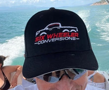 Load image into Gallery viewer, A person wearing a black baseball cap with the text 'SIX WHEELER CONVERSIONS' printed on the front, with a background of a boat on water.